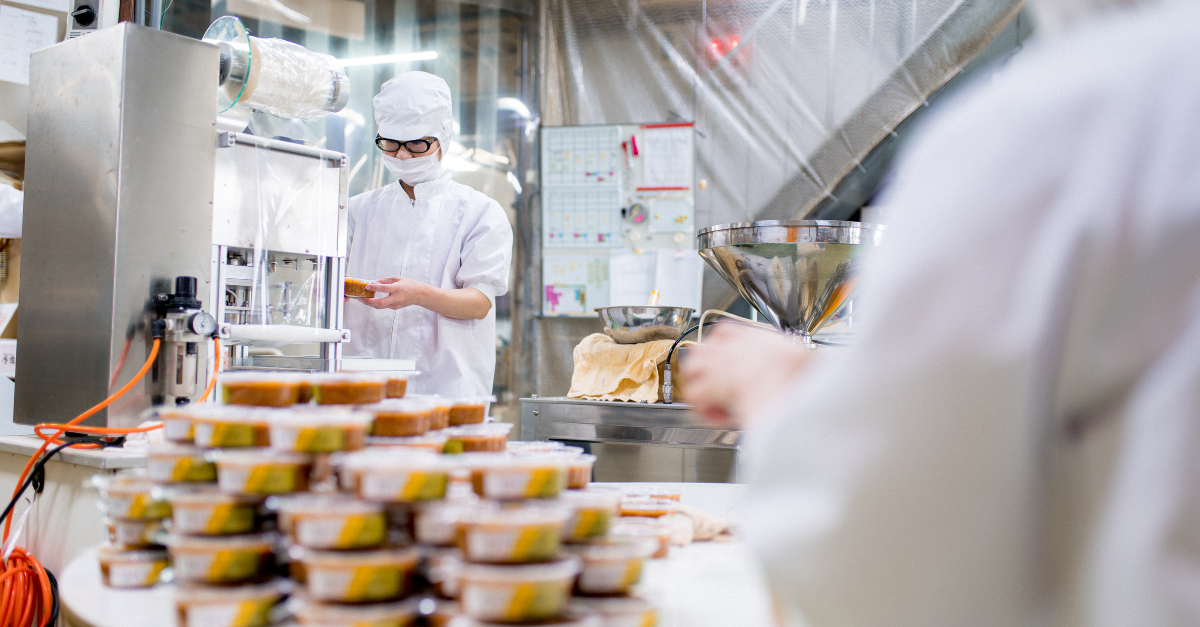 Pessoa com bata de laboratório e máscara a colocar tampas em embalagens de alimentos atrás de um monte de embalagens